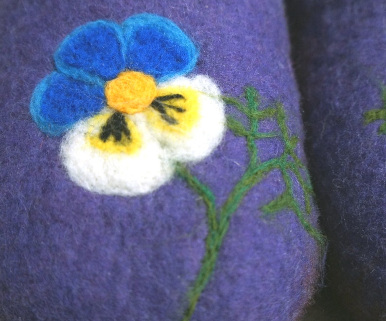 May include: A deep purple felted wool slipper featuring a needle-felted pansy flower design. The flower has bright blue upper petals, white lower petals with yellow centers, and dark markings. A thin green stem is stitched onto the purple surface.