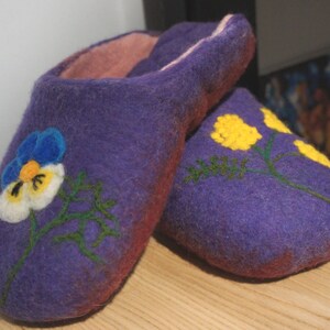 May include: A pair of purple felted wool slippers with a soft pink interior lining. The left slipper features a blue, white, and yellow pansy flower design, while the right slipper displays yellow floral sprigs with green stems and leaves.