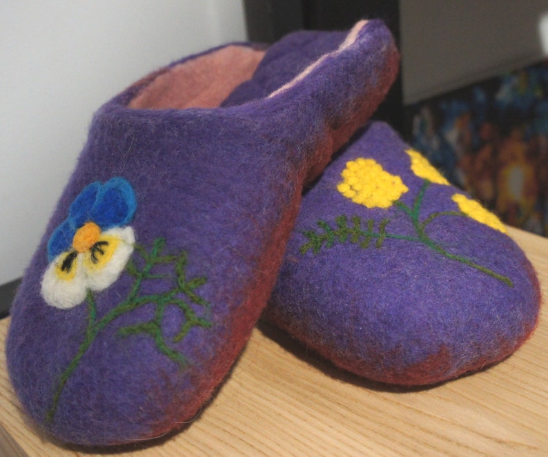 May include: A pair of purple felted wool slippers with a soft pink interior lining. The left slipper features a blue, white, and yellow pansy flower design, while the right slipper displays yellow floral sprigs with green stems and leaves.