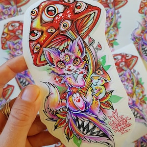 Op de afbeelding: Een sticker met een kleurrijke, cartoonachtige illustratie van een kat met meerdere ogen, die onder een rode paddenstoel met veel ogen zit. De paddenstoel heeft een witte steel en is omringd door groene bladeren. De kat draagt een roze halsband met een hartvormige hanger. De sticker heeft de tekst "My Cute Creatures" in zwart en wit.