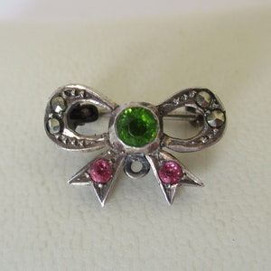 May include: A silver bow brooch with a green gemstone in the centre and two pink gemstones on the sides.