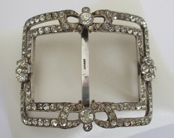 May include: A silver-toned belt buckle with a square frame and a decorative bow design. The buckle is adorned with clear rhinestones and has the word "Argent" engraved on the back.