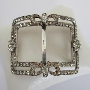 May include: A silver-toned belt buckle with a square frame and a decorative bow design. The buckle is adorned with clear rhinestones and has the word "Argent" engraved on the back.