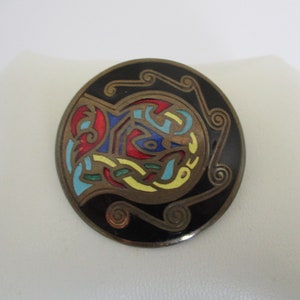 May include: A round, black enamel pin with a colorful Celtic knot design. The knot is in shades of red, blue, yellow, and green.