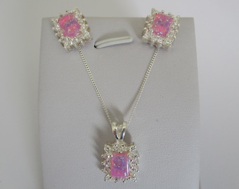 May include: A jewelry set featuring a pendant necklace and matching earrings. Each piece has a rectangular, pink opal center stone surrounded by small, clear crystals. The set is displayed on a gray stand.