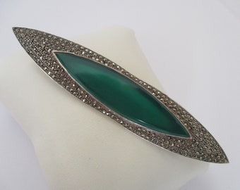 May include: A silver brooch with a green stone set in a marcasite frame. The brooch is shaped like a long oval.