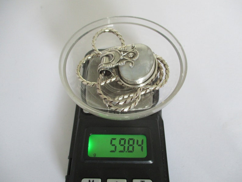 May include: A silver chain necklace with a silver pendant featuring a white stone and intricate details. The necklace and pendant are on a digital scale that reads 59.84.
