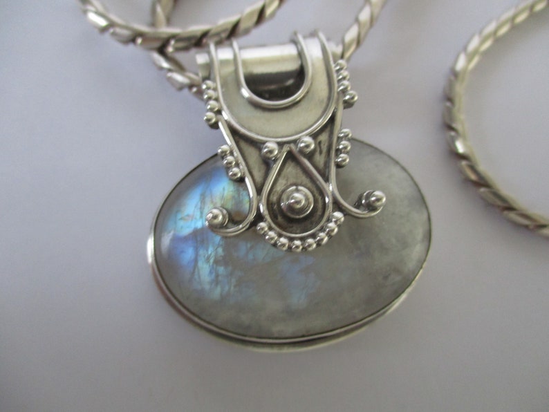 May include: A silver pendant with a large oval moonstone set in a silver bezel. The pendant has a detailed silver bail with a decorative design.