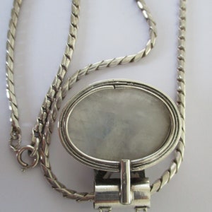 May include: A silver chain necklace with a silver oval locket pendant. The locket has a smooth, white interior and a detailed design around the edge. The chain is a twisted rope style.