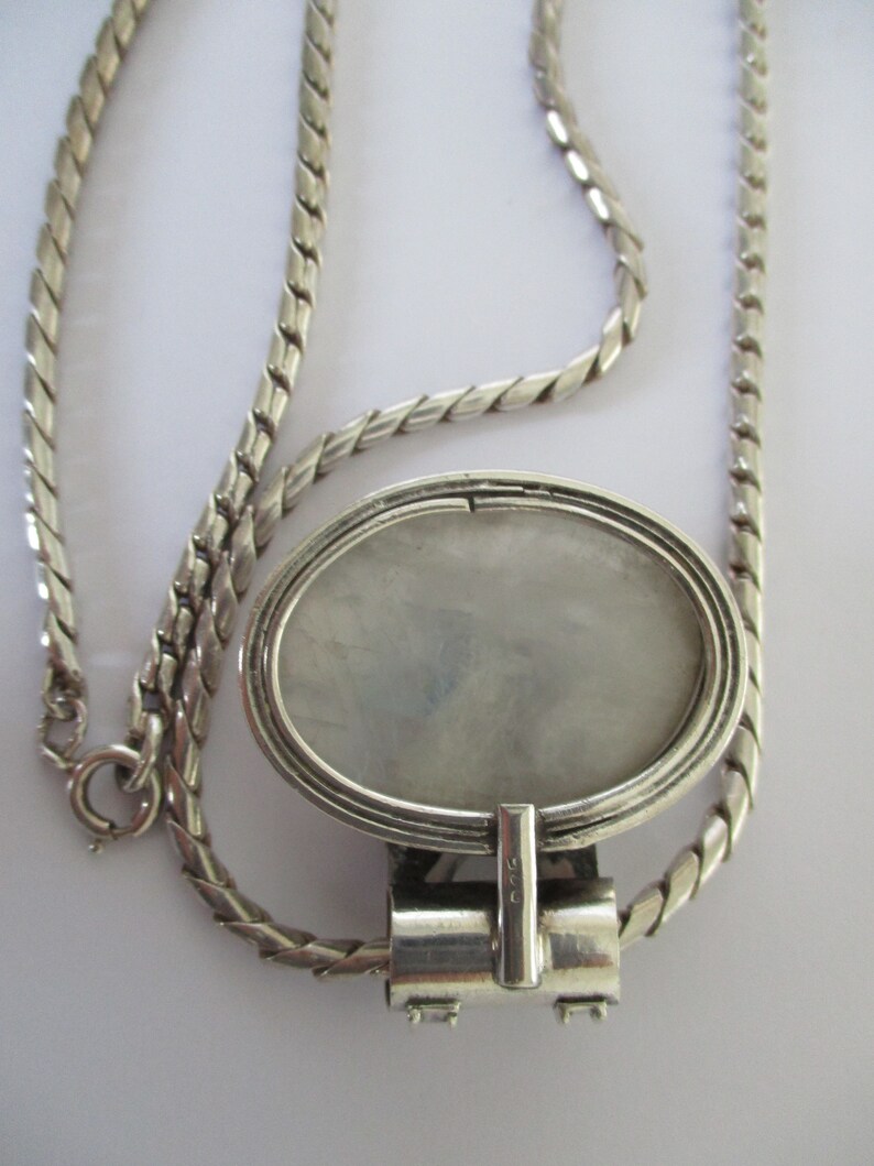 May include: A silver chain necklace with a silver oval locket pendant. The locket has a smooth, white interior and a detailed design around the edge. The chain is a twisted rope style.