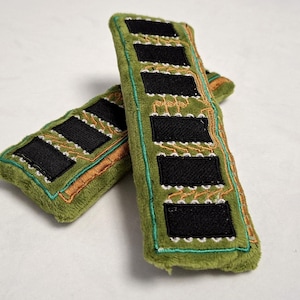 May include: Two plush rectangular items in olive green fabric, designed to resemble computer RAM sticks. Each features embroidered black rectangular patches and orange and teal circuit board patterns stitched onto the soft, fuzzy surface.