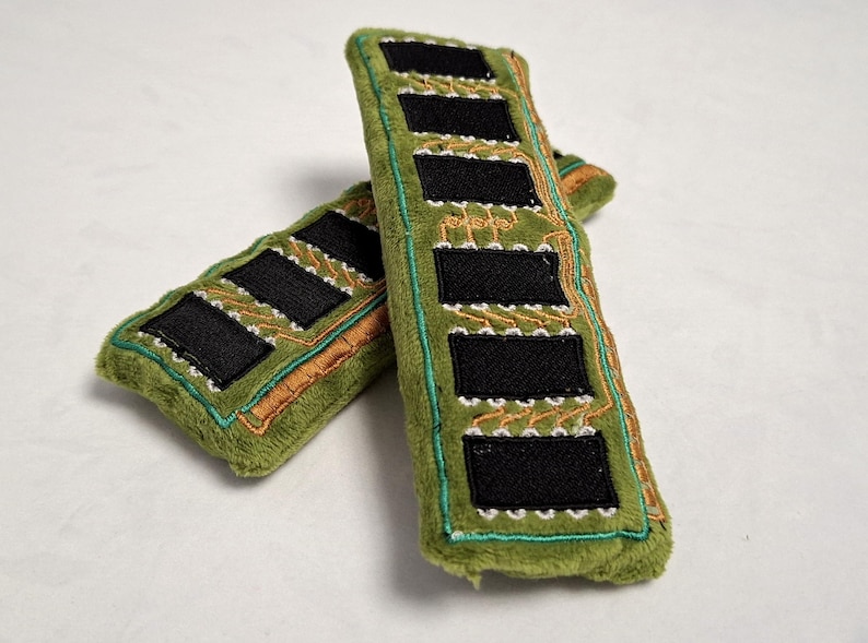 May include: Two plush rectangular items in olive green fabric, designed to resemble computer RAM sticks. Each features embroidered black rectangular patches and orange and teal circuit board patterns stitched onto the soft, fuzzy surface.