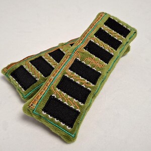 May include: Two rectangular plush items in olive green fabric, each featuring embroidered black rectangular patches arranged in a row. The design includes intricate gold and white thread detailing that mimics the appearance of a circuit board.
