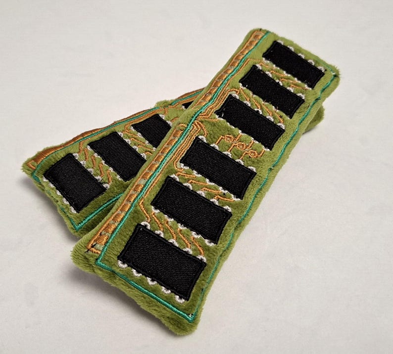 May include: Two rectangular plush items in olive green fabric, each featuring embroidered black rectangular patches arranged in a row. The design includes intricate gold and white thread detailing that mimics the appearance of a circuit board.