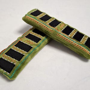May include: Two rectangular plush items in olive green, designed to resemble computer RAM sticks. Each piece features black felt rectangles arranged in a row, accented with gold and teal embroidered circuit board patterns on the soft fabric.