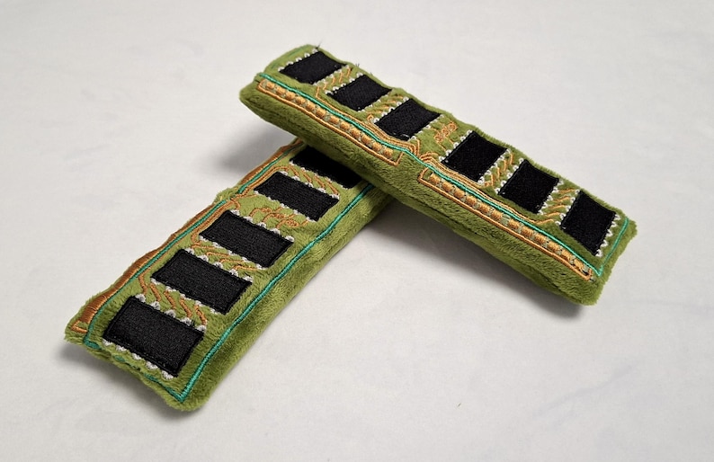 May include: Two rectangular plush items in olive green, designed to resemble computer RAM sticks. Each piece features black felt rectangles arranged in a row, accented with gold and teal embroidered circuit board patterns on the soft fabric.