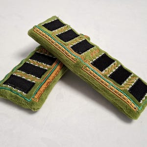 May include: Two plush rectangular items in olive green fabric, designed to resemble computer RAM sticks. Each features black felt squares, gold-colored embroidered circuit board patterns, and teal stitching along the edges of the soft material.