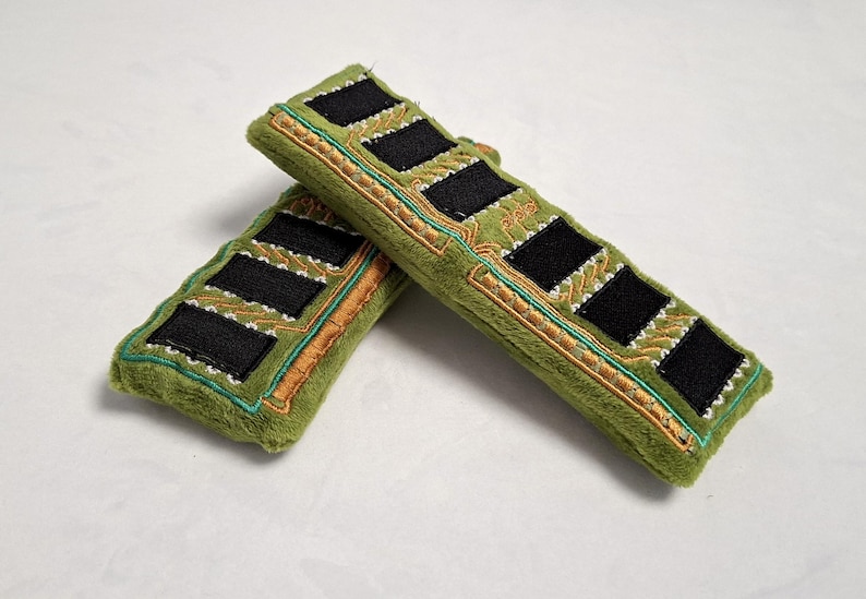 May include: Two plush rectangular items in olive green fabric, designed to resemble computer RAM sticks. Each features black felt squares, gold-colored embroidered circuit board patterns, and teal stitching along the edges of the soft material.