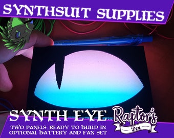 May include: Two black panels with a blue, pink, and white gradient eye design. The panels are for building a synth eye, and the text "SYNTH EYE Raptor's Den" is printed below the eye. The text "TWO PANELS, READY TO BUILD IN OPTIONAL BATTERY AND FAN SET" is printed below the logo.
