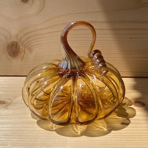 May include: A decorative glass pumpkin in shades of amber and gold. The pumpkin has a ribbed design and a curved stem. The glass pumpkin is sitting on a wooden surface and is casting a shadow.