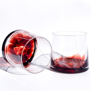 May include: Two clear, lowball glasses with a dark red base and a thin, white, linear design. One glass is upright, the other is tilted on its side. The glasses are on a white surface, casting shadows.