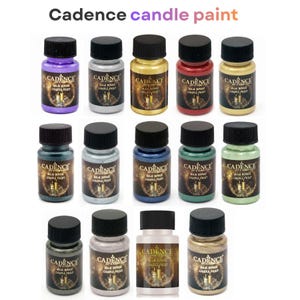 Peut inclure: Une collection de petits flacons de peinture de différentes couleurs, dont le violet, l'argent, l'or et le rouge. Chaque flacon a un bouchon noir et une étiquette indiquant "Cadence candle paint". Les flacons sont disposés en rangées.