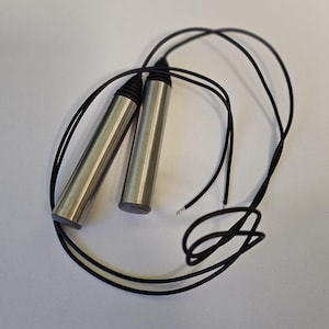 Puede incluir: Dos objetos cilíndricos de acero inoxidable con cables negros adjuntos. Los cilindros están uno al lado del otro, con los cables que los rodean. Los extremos de los cilindros son de un gris más oscuro. Los cables son negros y parecen aislados.