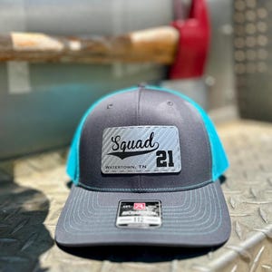 May include: A gray and teal baseball cap with the word "Squad" and the number "21" on a patch. The text "Watertown, TN" is below the word "Squad". The hat has a structured front and a mesh back.