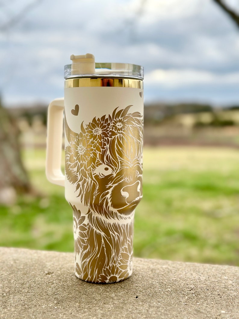 Peut inclure: Mug de voyage blanc avec un motif dor&eacute; de vache Highland avec des fleurs. Le mug a un couvercle et une poign&eacute;e.