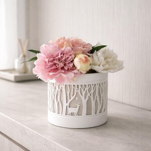 Könnte beinhalten: Eine weiße, zylindrische Vase mit einem ausgeschnittenen Waldmotiv und einer Hirschsilhouette, gefüllt mit rosa, pfirsichfarbenen und weißen Pfingstrosen. Die Vase steht auf einer hellen Oberfläche und schafft eine ruhige, von der Natur inspirierte Präsentation.