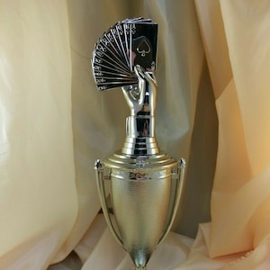 May include: A silver trophy with a hand holding a fanned deck of playing cards, featuring the Ace of Spades. The trophy has a gold-colored cup and a black and green marble base. A unique award for card game enthusiasts.