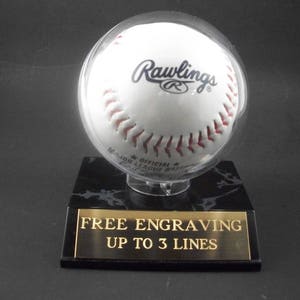 May include: A white Rawlings Major League baseball in a clear display case, resting on a black marble base with a gold plaque. The plaque reads "FREE ENGRAVING UP TO 3 LINES". The baseball has red stitching.