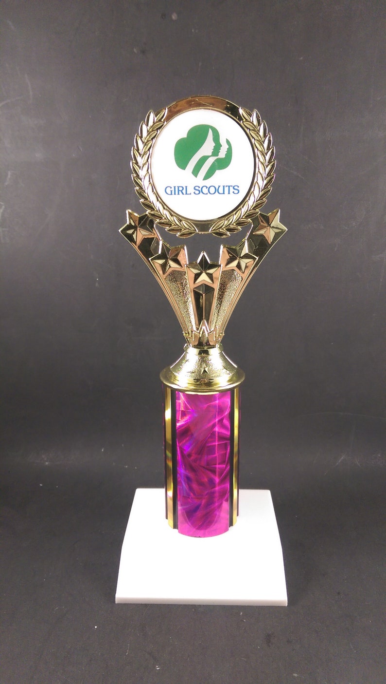 May include: A gold trophy with a pink base and a Girl Scouts logo on the top. The trophy has five gold stars around the base of the logo.