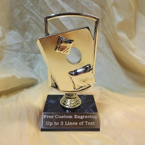 May include: A gold-toned trophy featuring a central rectangular plate with two embossed pillow shapes and a circular cutout, framed by a metallic arch. The trophy stands on a black marble-patterned base with a silver plaque reading Free Custom Engraving Up to 3 Lines of Text.
