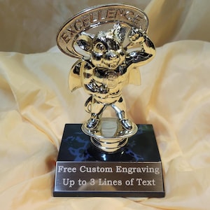 May include: A gold-colored trophy featuring a cartoon character with a superhero pose, standing on a round base atop a black and blue marble-patterned base. The trophy has a banner that says "EXCELLENCE". The base has text that says "Free Custom Engraving Up to 3 Lines of Text".