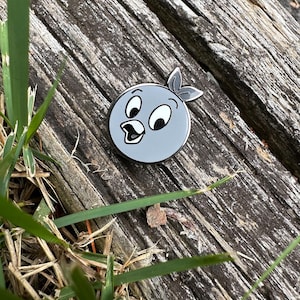 Könnte beinhalten: Runder, grauer Emaille-Pin mit einem Cartoon-Vogel mit weißem Schnabel, Augen und einem blattförmigen Detail. Der Pin liegt auf einer verwitterten Holzoberfläche mit grünen Grashalmen im Vordergrund.