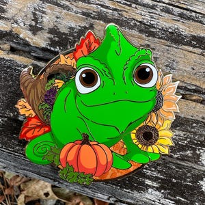 May include: A green chameleon enamel pin with large brown eyes, surrounded by a harvest-themed design. The pin features a pumpkin, sunflower, and a cornucopia filled with grapes and leaves. The pin is set against a wooden background.