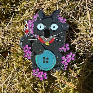 May include: A whimsical enamel pin featuring a black cat with large blue eyes, a red collar with a gold bell, and a large turquoise button. The cat is surrounded by purple flowers and a small red and white character.