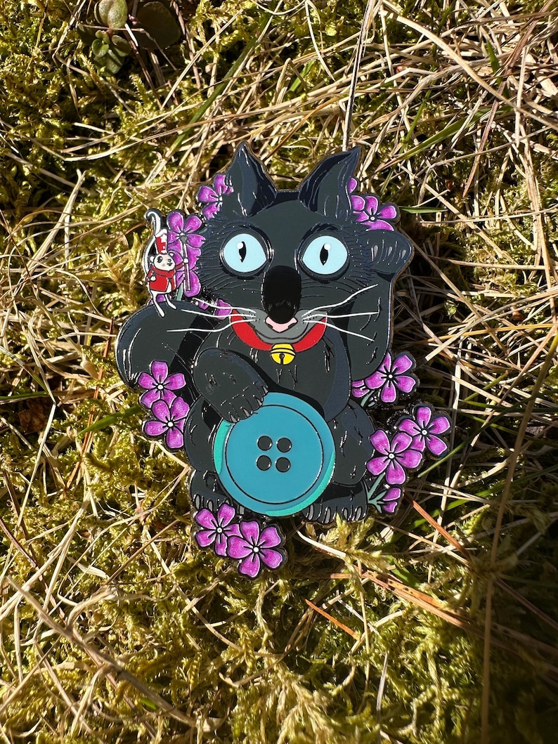 May include: A whimsical enamel pin featuring a black cat with large blue eyes, a red collar with a gold bell, and a large turquoise button. The cat is surrounded by purple flowers and a small red and white character.