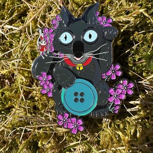 May include: A black cat enamel pin with a turquoise button, purple flowers, and a red collar with a gold bell. A small red and white rabbit pin is attached to the cat. The pin is set against a background of green moss and brown grass.