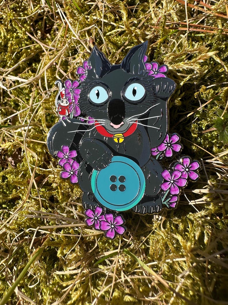 May include: A black cat enamel pin with a turquoise button, purple flowers, and a red collar with a gold bell. A small red and white rabbit pin is attached to the cat. The pin is set against a background of green moss and brown grass.