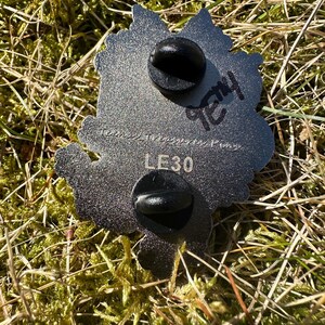 May include: A silver-colored metal pin with two black pin backs. The pin has a leaf-like shape with a textured surface. The text "LE30" is visible, along with handwritten text. The pin is set against a background of green moss and grass.