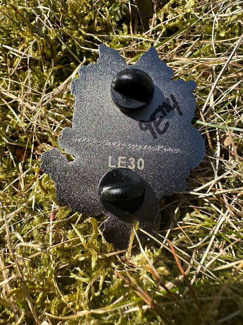 May include: A silver-colored metal pin with two black pin backs. The pin has a leaf-like shape with a textured surface. The text "LE30" is visible, along with handwritten text. The pin is set against a background of green moss and grass.