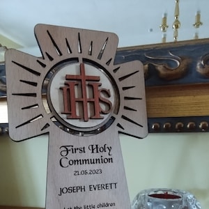 May include: A wooden cross with a sunburst design and a round center featuring the letters "IHS" in red. The cross is engraved with the text "First Holy Communion", the date "21.05.2023", the name "JOSEPH EVERETT", a quote from Matthew 19:14, and the name "St Norberts Spalding".
