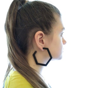 May include: A person wearing large black hexagonal hoop earrings.