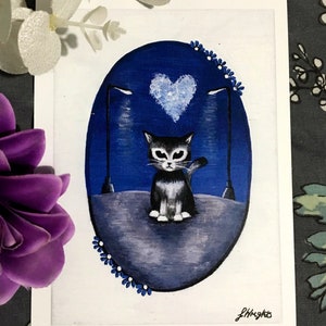 May include: An original painting of a black and white cat sitting on a grey surface, with a blue heart above its head. The cat is in an oval shape with a dark blue background, two street lamps, and small blue flowers around the edge.