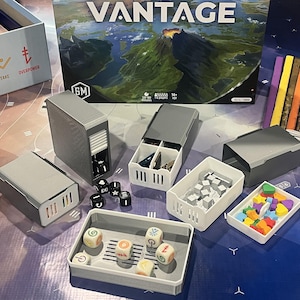 May include: A board game called "VANTAGE" with game components. The image shows grey storage containers, dice, and game pieces in white and colourful trays. The game box is visible in the background.