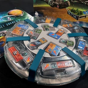 May include: A clear plastic game component with a white center and teal straps. The center has multiple slots that hold small, colorful cards with images of planets and spaceships. The text "Planet Unknown" is visible on a nearby game box.