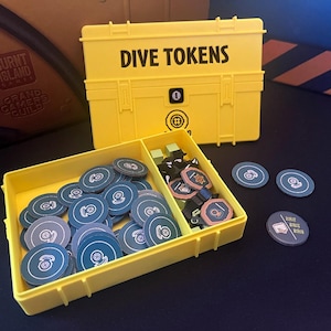 May include: A yellow plastic box with the text "Dive Tokens" on the lid. The box is open and contains a variety of black and white tokens, as well as some green and orange tokens. The box is sitting on a black surface.