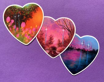 Puede incluir: Tres pegatinas en forma de corazón con pinturas de paisajes. Una presenta tulipanes rosas y un cielo naranja vibrante. Otra representa una puesta de sol rosa y púrpura sobre el agua. La tercera muestra una puesta de sol azul y rosa.